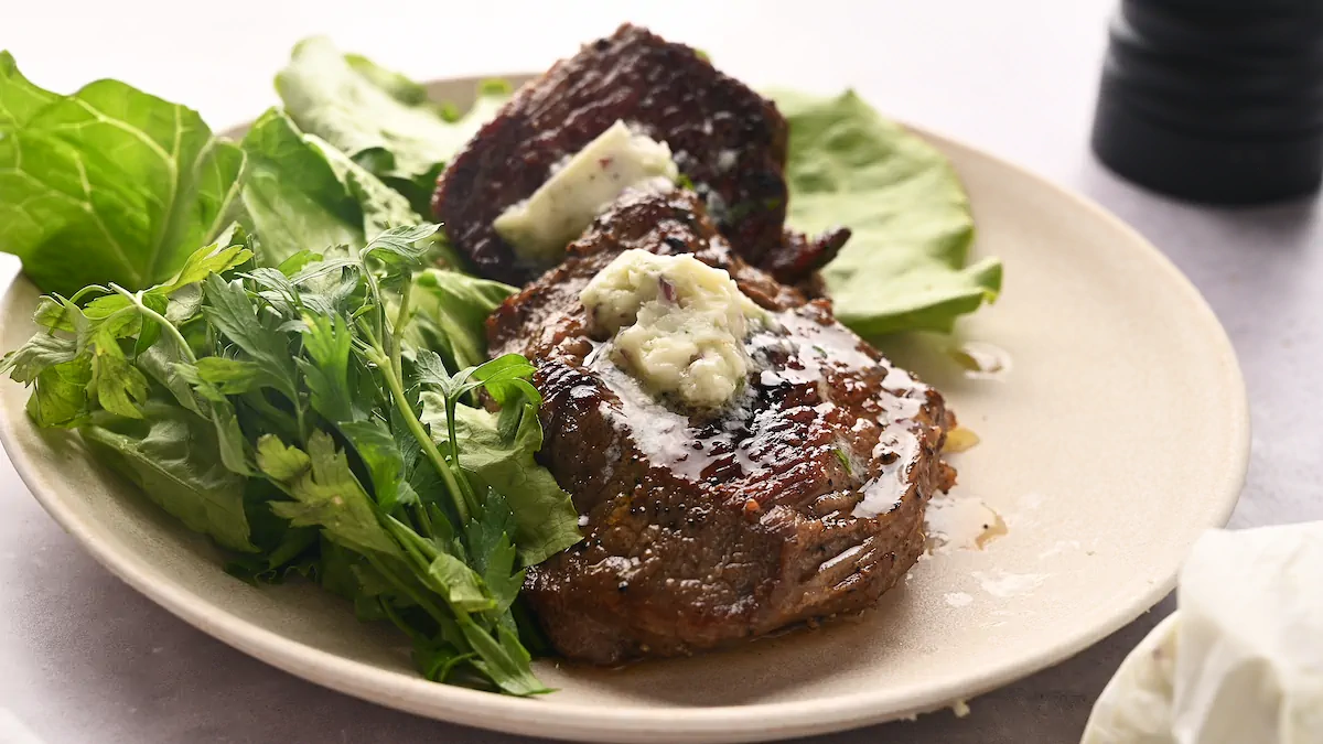 Herb butter-topped flat iron steak served on a plate with a side of fresh greens.