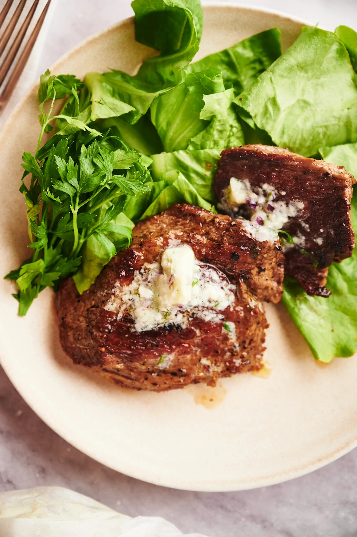 Homemade flat iron steak topped with herb butter and served alongside fresh greens on a plate.