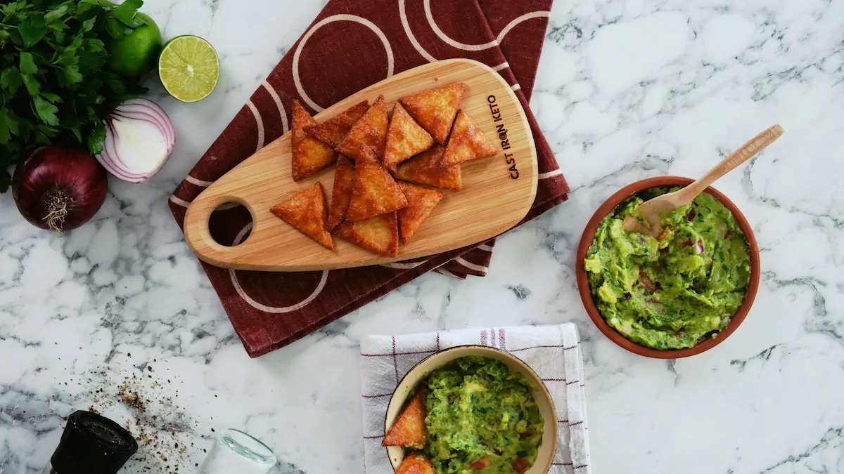Keto tortilla chips on a wooden board alongside a bowl of guacamole dip.