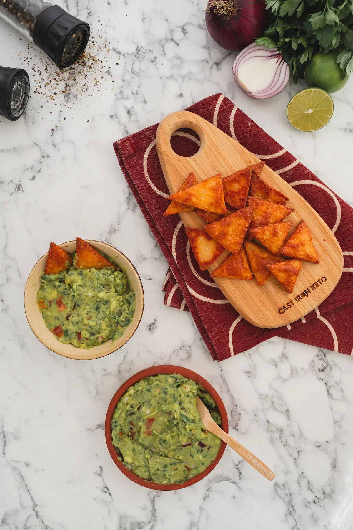 Keto-friendly tortilla chips served on a wooden board beside two guacamole bowls.