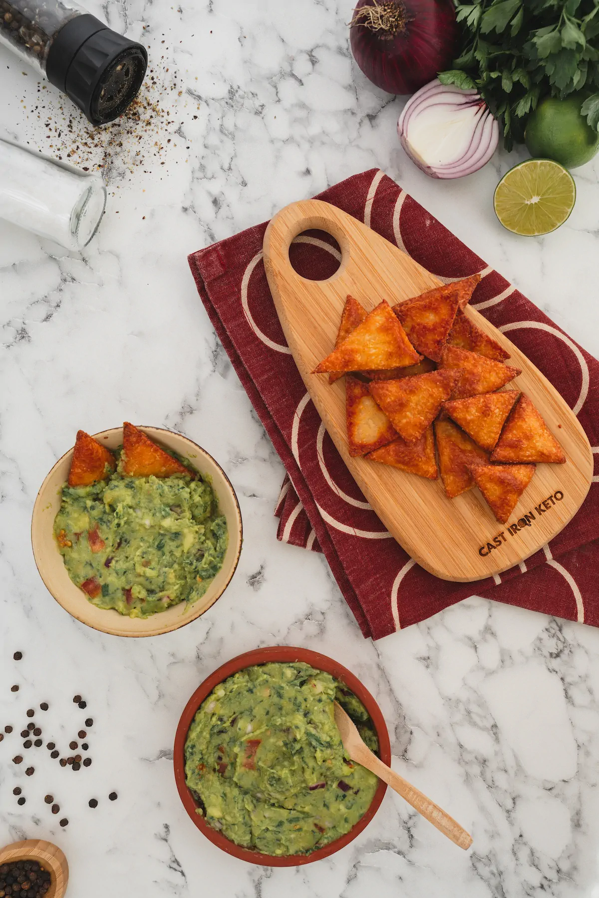 Keto tortilla chips on a wooden board next to two bowls of guacamole.