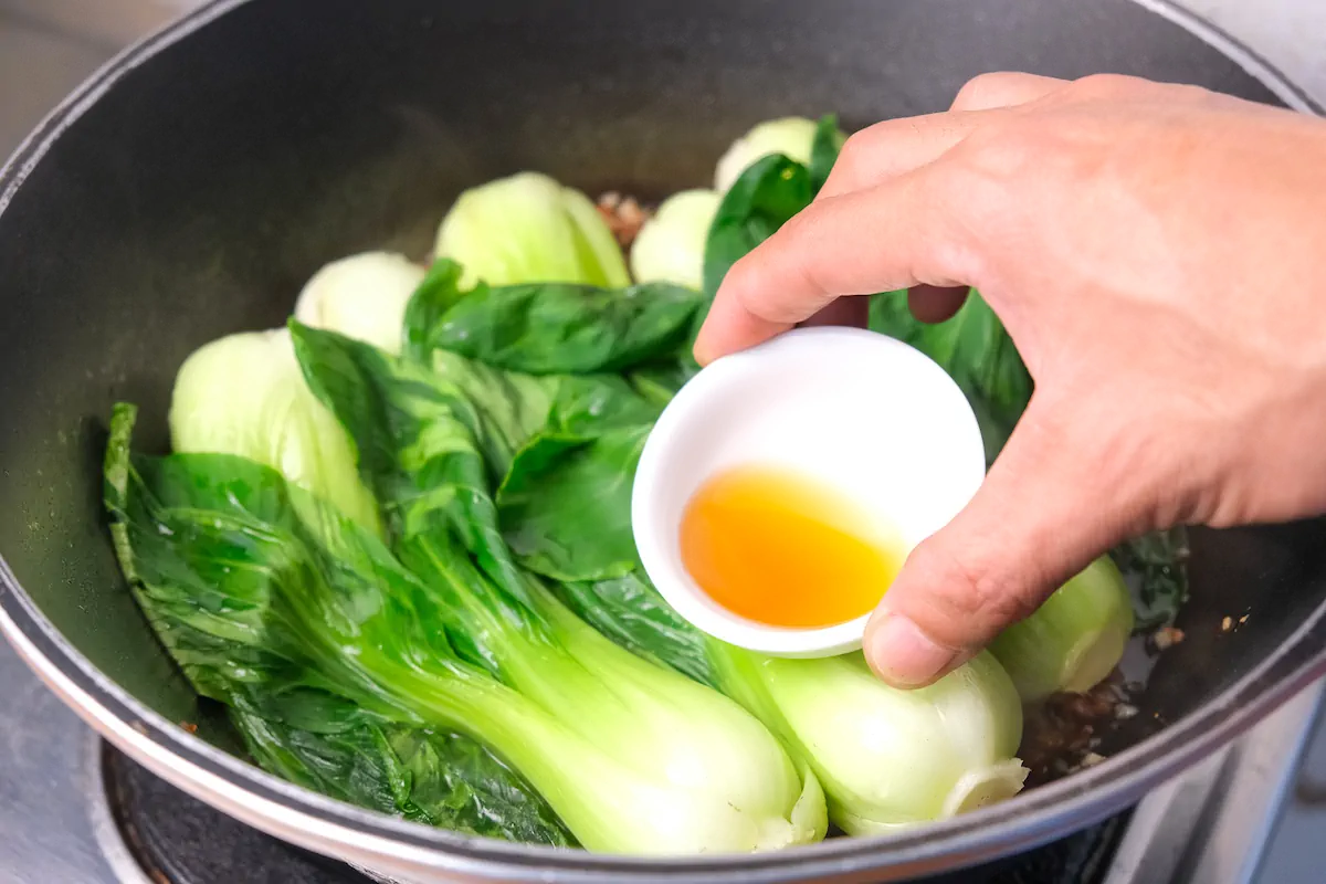 About to pour sesame oil from a bowl to the pan with Chinese style bok choy.