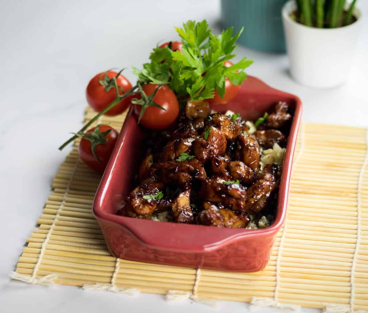 Keto bourbon chicken on a bed of cauliflower rice in a red serving dish, garnished with cilantro and red tomatoes.