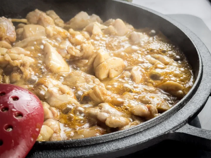 Chicken simmering with bourbon sauce in a cast iron skillet, with a red spatula resting on the pan.