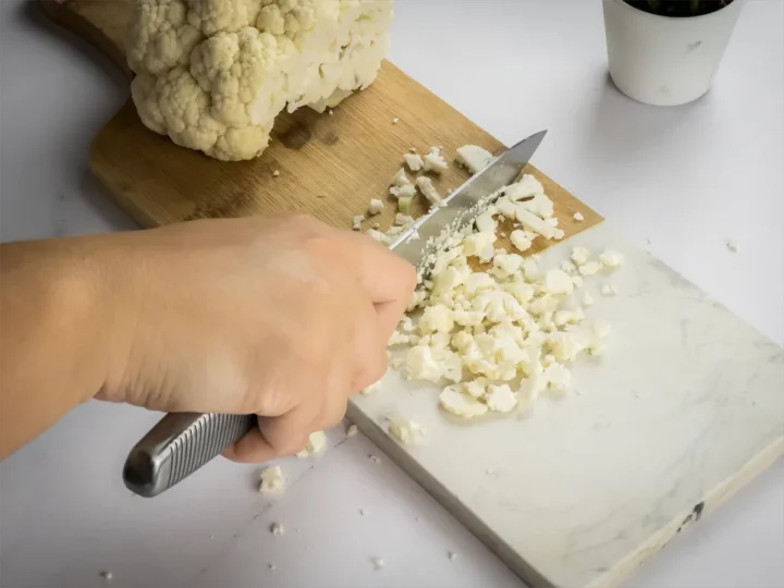 A hand is chopping cauliflower into small pieces on a wooden chopping board.