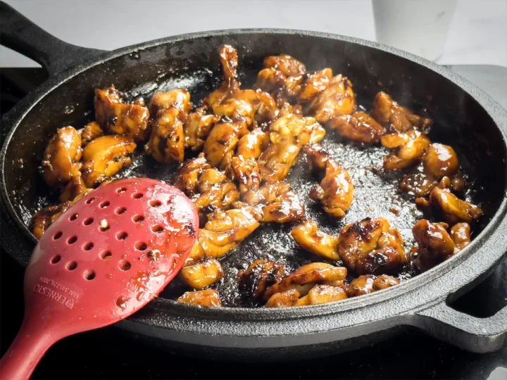 Chicken pieces glazed with bourbon sauce in the cast iron skillet, with a red spatula resting on the pan.