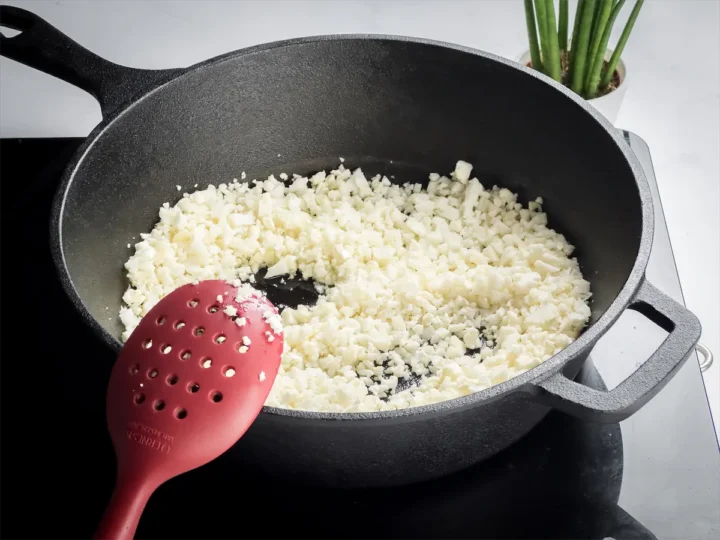 Chopped cauliflower is cooking in the cast iron skillet with oil while a red spatula rests on the skillet.