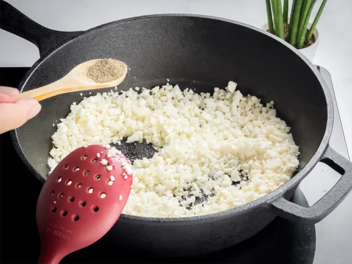 A hand holding a spoonful of black pepper over cauliflower rice cooking in a cast iron skillet.