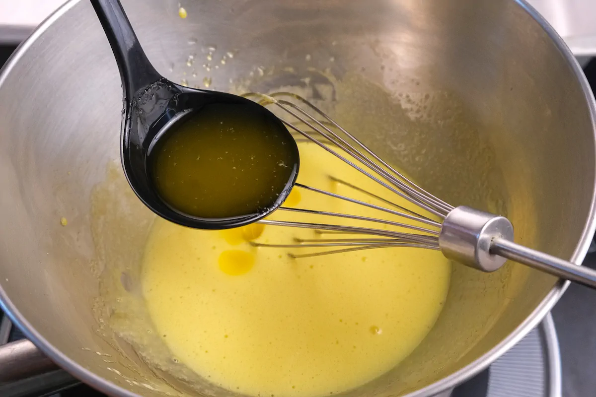 Melted butter is ready to be poured from a ladle into the mixture of egg yolk and lemon juice in a mixing bowl.