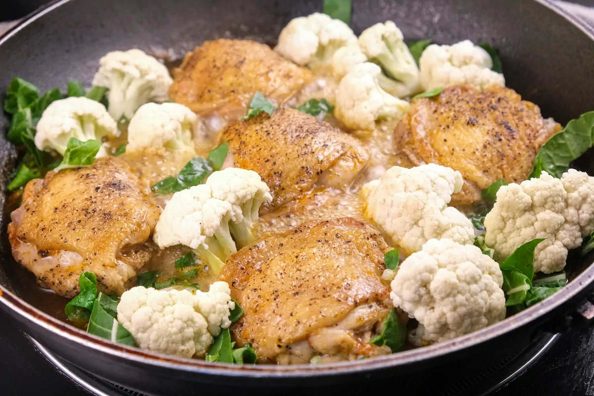 Adding cauliflower and spinach to the cast iron skillet with chicken thighs.
