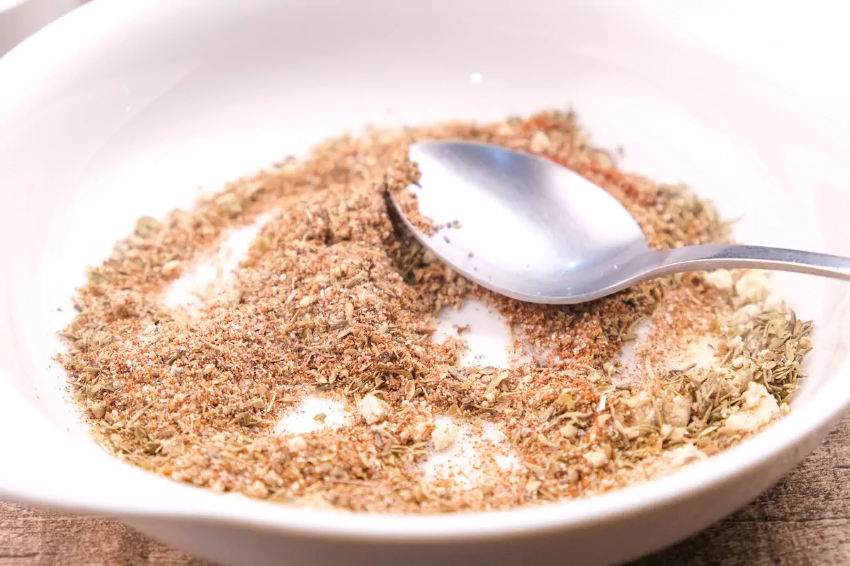 Combining sea salt, black pepper, garlic powder, onion powder, cumin, paprika, dried oregano, and thyme with a spoon for the dry rub.