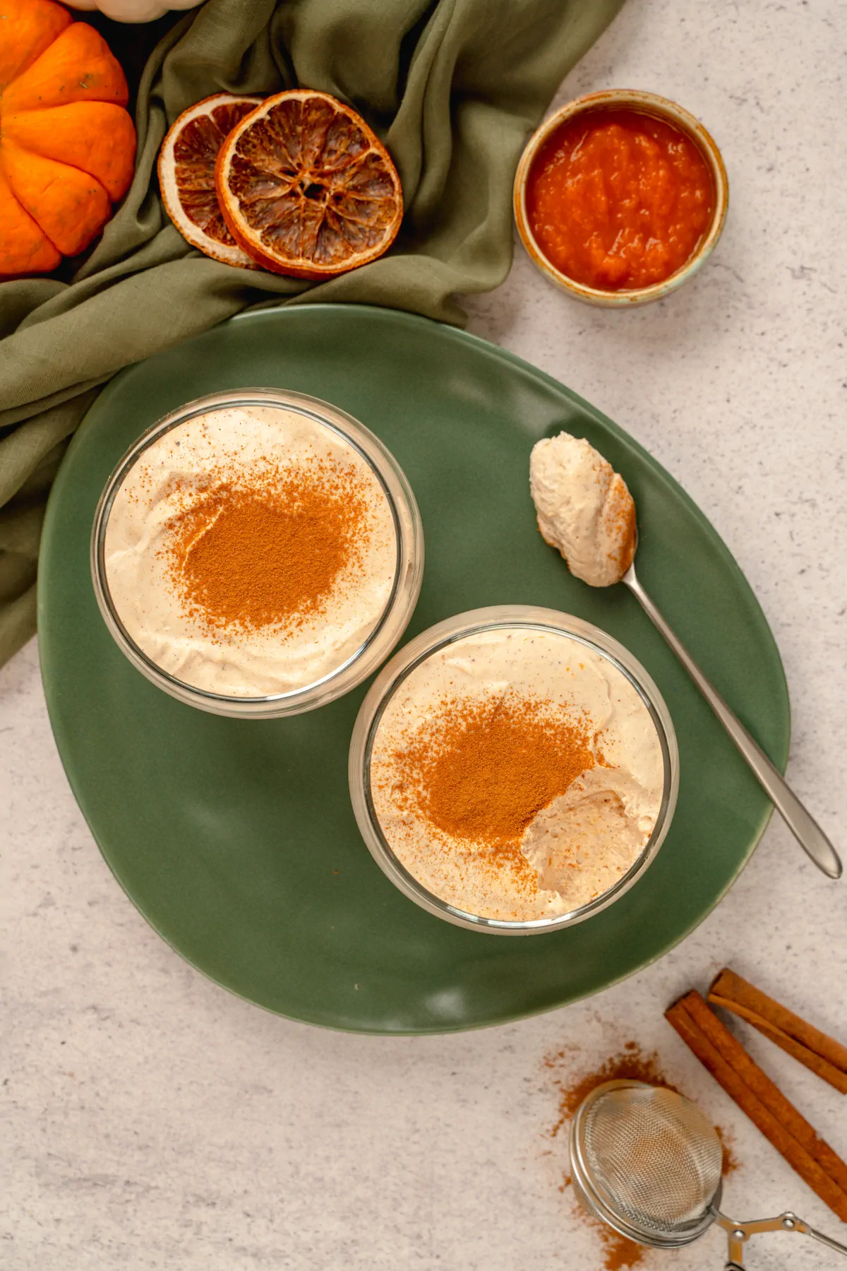 Homemade keto pumpkin cheesecake mousse served in two transparent glasses dusted with cinnamon powder, where some portions from one of the glasses and a spoonful of the same dessert placed on a plate.