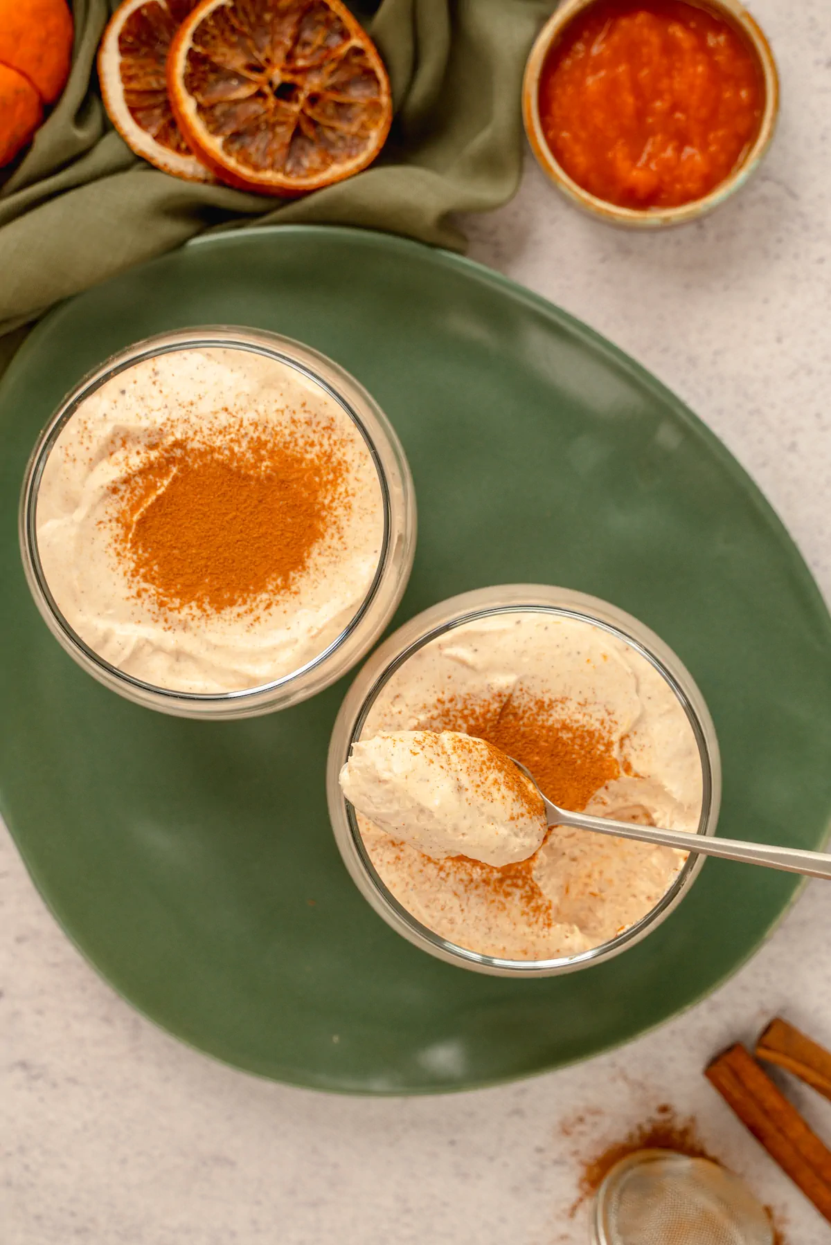 Keto pumpkin cheesecake mousse served in two transparent glasses garnished with cinnamon powder, where one glass remains untouched, while some portions is eaten from the other glass and a spoonful is ready to be eaten too.