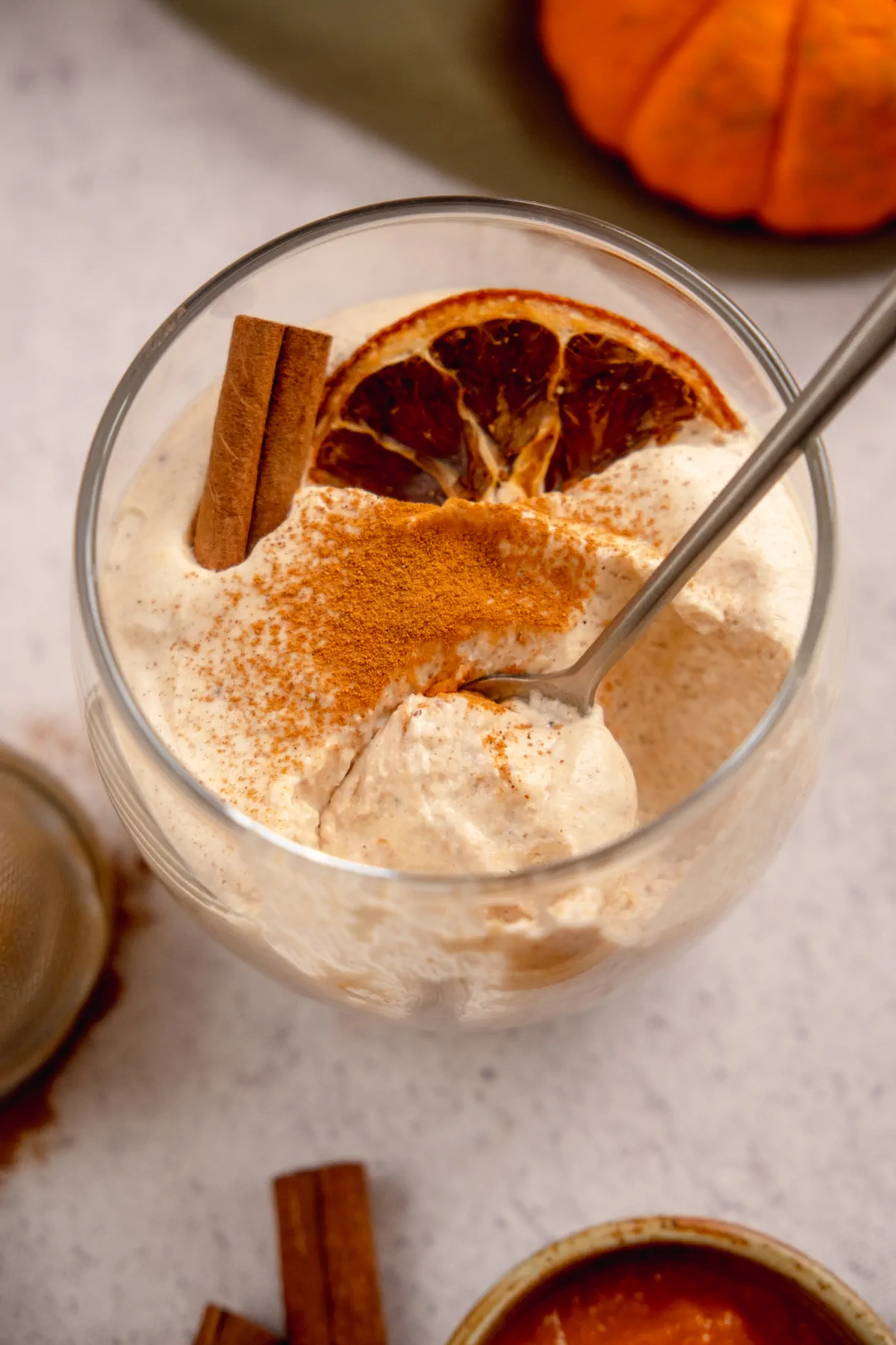 A focused photo of homemade keto pumpkin cheesecake mousse getting eaten with a spoon from a transparent glass garnished with cinnamon powder, a dried orange slice and cinnamon stick.
