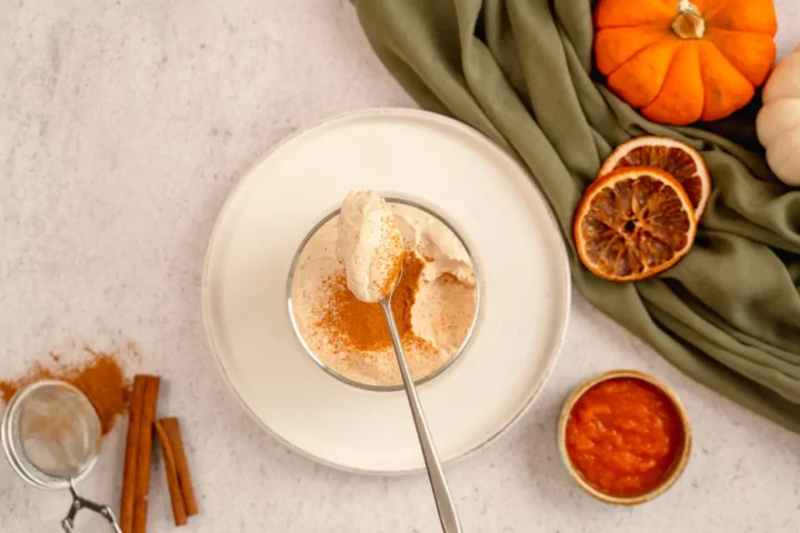 Homemade keto pumpkin cheesecake mousse served in a transparent glass dusted with cinnamon powder, with a spoonful of the same dessert placed on the glass alongside a small bowl of pumpkin puree.
