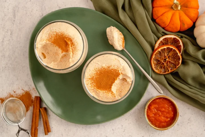 Keto pumpkin cheesecake mousse served in two transparent glasses dusted with cinnamon powder, where some portions from each eaten and a spoonful of the same dessert placed on a plate.