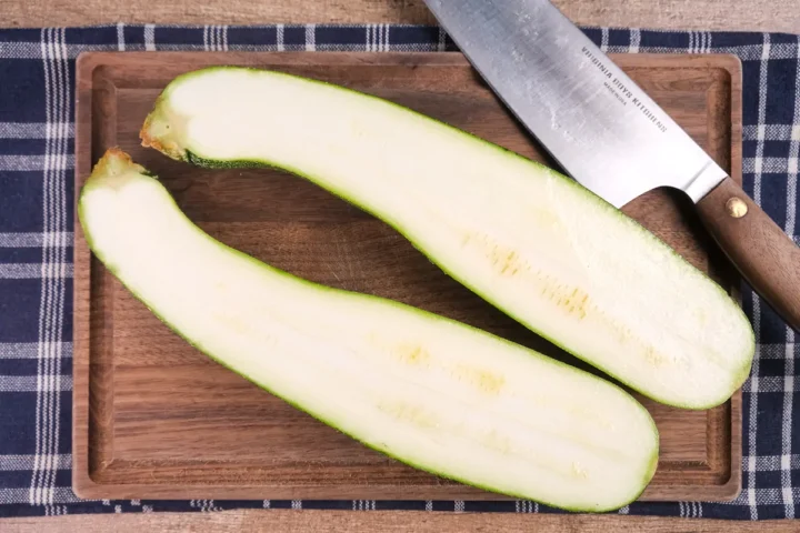 Lengthwise halved zucchini on a wooden chopping board with a knife.