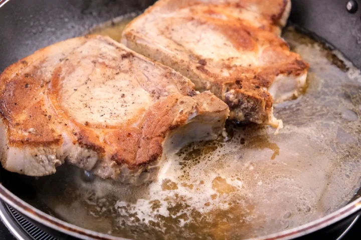 Adding beef broth to the pan with golden bone-in pork chops.