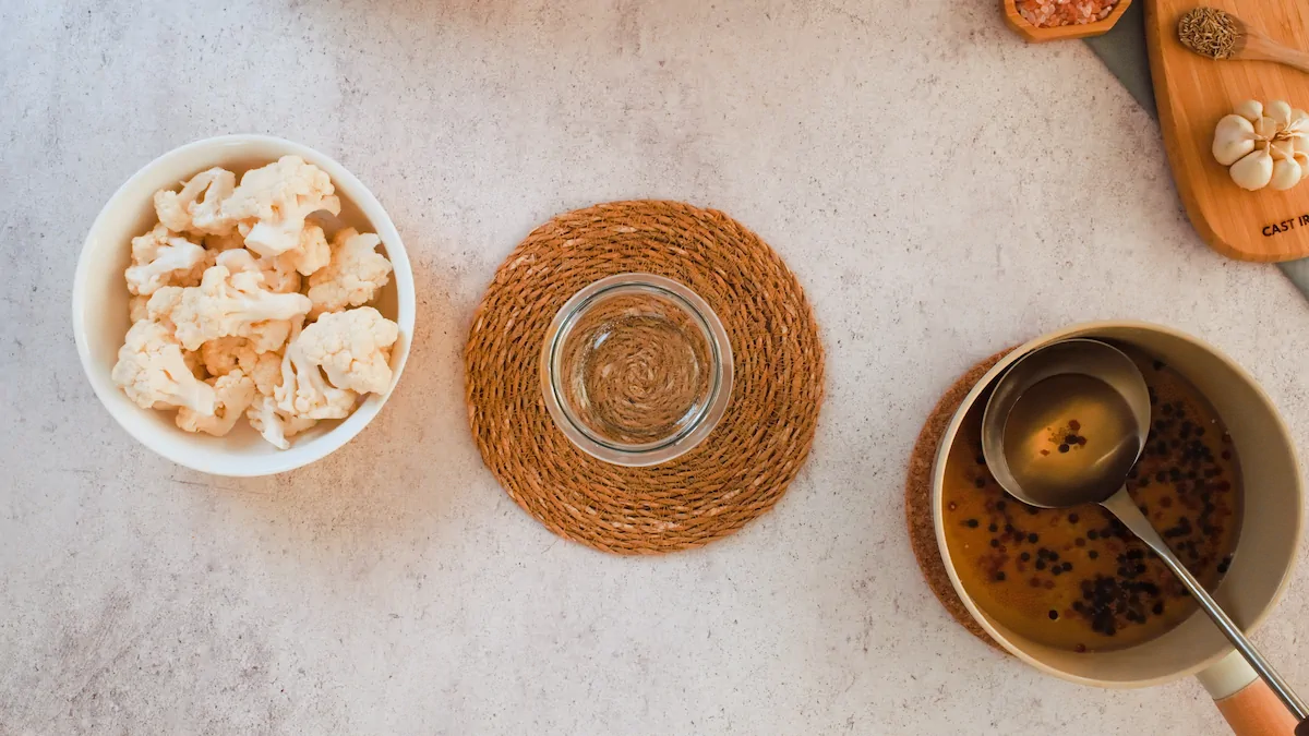 Cauliflower florets, empty glass jar and brine with a ladle arranged next to each other.