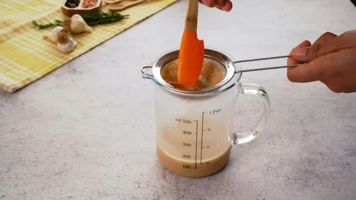 A hand holding a fine strainer over a glass measuring cup with keto gravy.