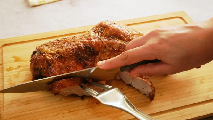 Slicing the roasted turkey leg with a knife and a fork on a chopping board.