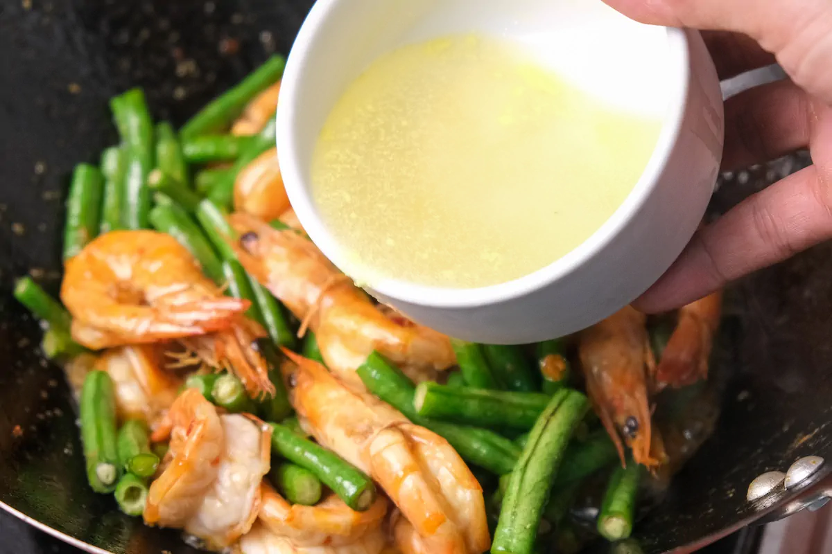 Chicken broth about to be poured from a white bowl to the wok with stir-fried shrimp and string beans.