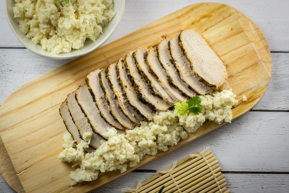 Top shot of slow-cooked pork loin and a side of mashed cauliflower on a wooden board.