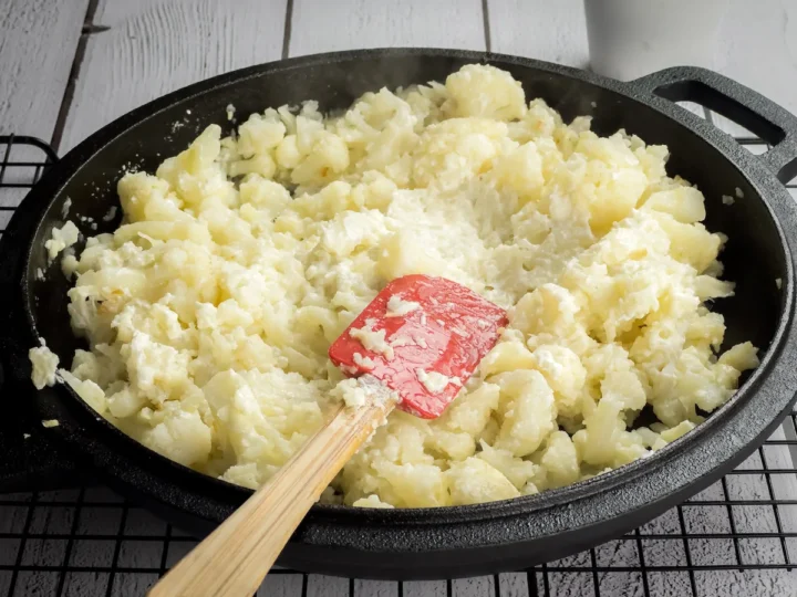 Mixing the cream cheese and grated Parmesan cheese into steamed cauliflower with a silicone spatula in a cast iron skillet.