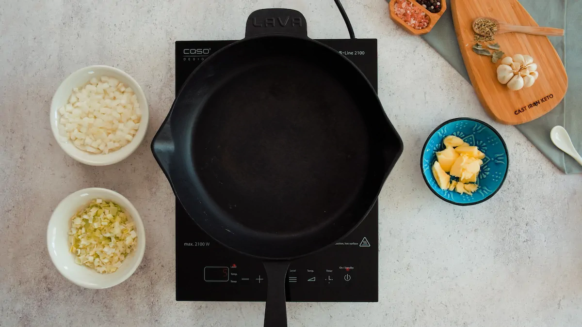 A cast iron skillet getting heated on an induction cooktop alongside butter, diced onion and diced leeks.