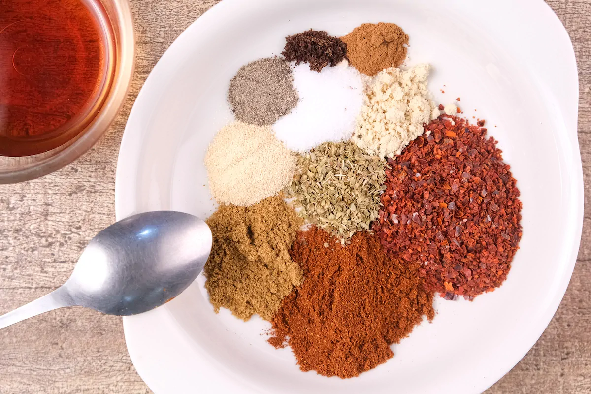 The required amount of spices including paprika, chili powder, dried oregano, cumin powder, granulated garlic, onion powder, sea salt, black pepper, ground cinnamon, and ground cloves are in a bowl.