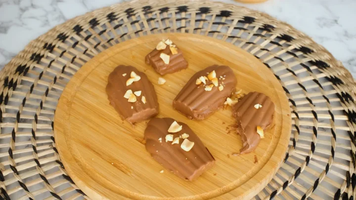 Keto chocolate fudge displayed on a round wooden board topped with roughly chopped roasted peanuts.