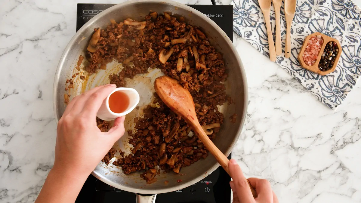 Pouring red wine vinegar to the ground beef and mushroom cooking in a stainless steel pan.