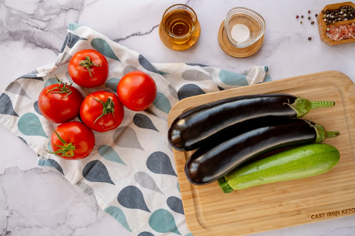 Tomatoes, egg plants and zucchini ready to be sliced for ratatouille alongside olive oil and salt.