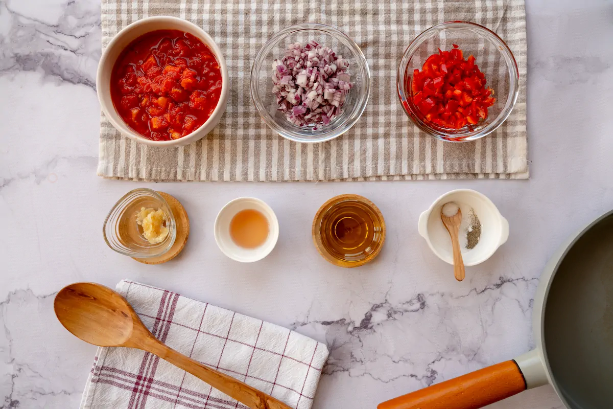 All the ingredients including olive oil, chopped tomatoes, diced onion, diced red bell pepper, minced garlic, apple cider vinegar, and sea salt to make ratatouille sauce gathered and arranged on the table.