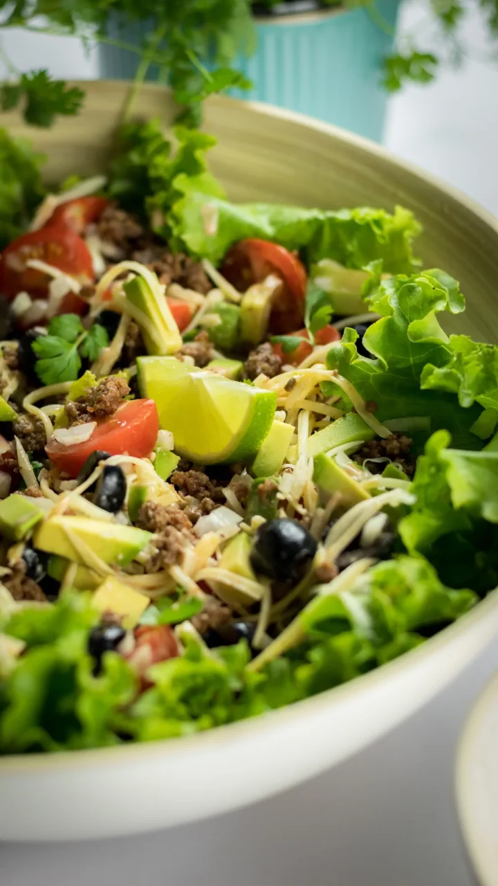 A close-up shot of a lemon wedge, black olives, and greens of a keto taco salad in a bowl.