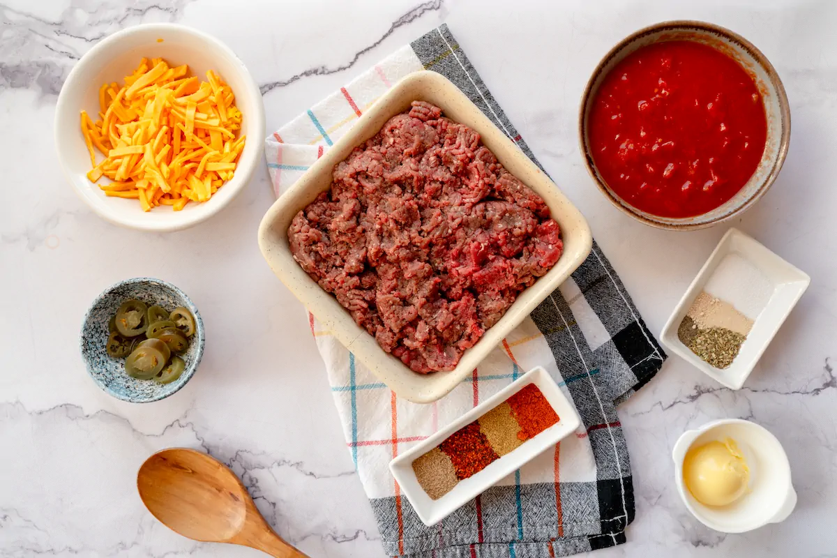 All the ingredients including ground beef, butter, Tex-Mex seasoning, diced tomatoes (canned), pickled jalapeños, and shredded cheddar cheese gathered and arranged on the table.