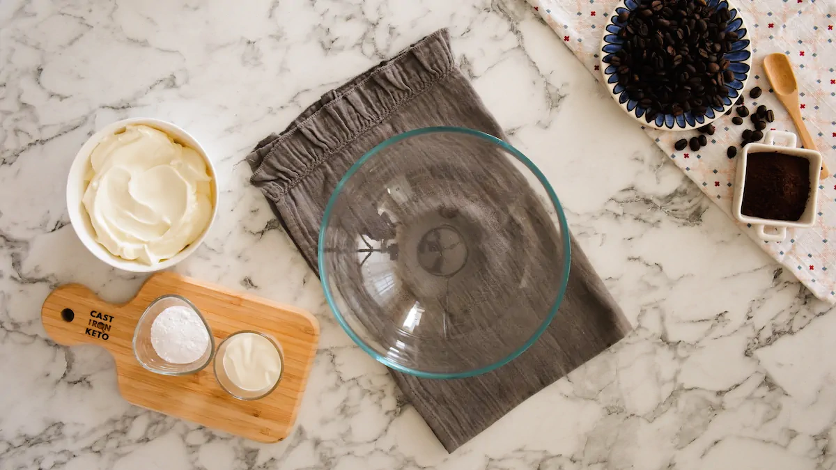 An empty mixing bowl alongside mascarpone cheese, heavy cream, and powdered erythritol.