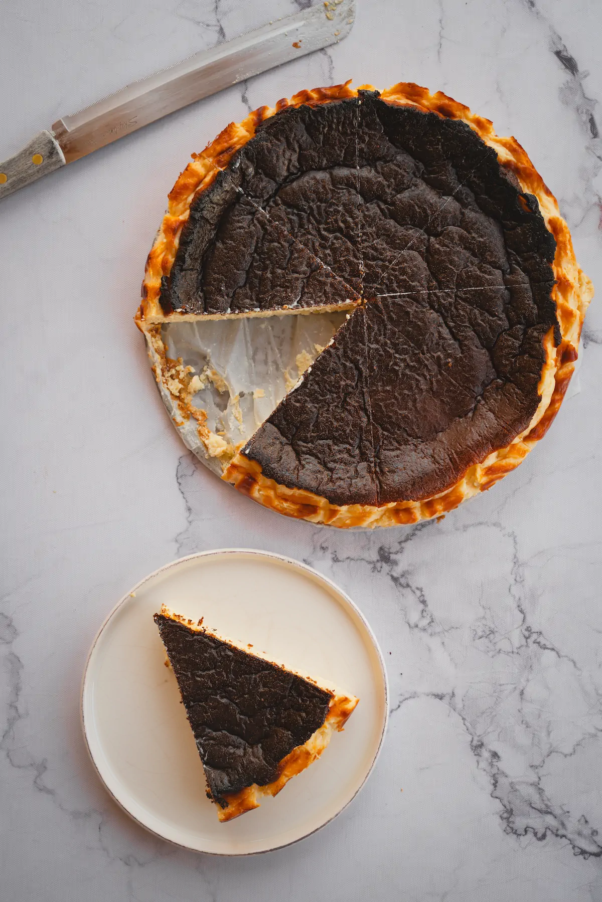 A plate holds a slice of burnt Basque cheesecake, cut from the whole cake.