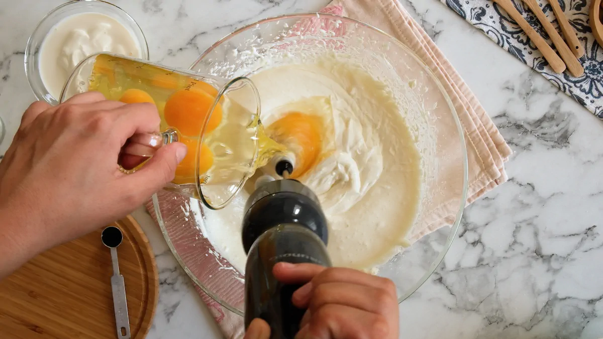 A hand is adding eggs to the cream cheese mixture, while the other hand is blending with a hand blender.