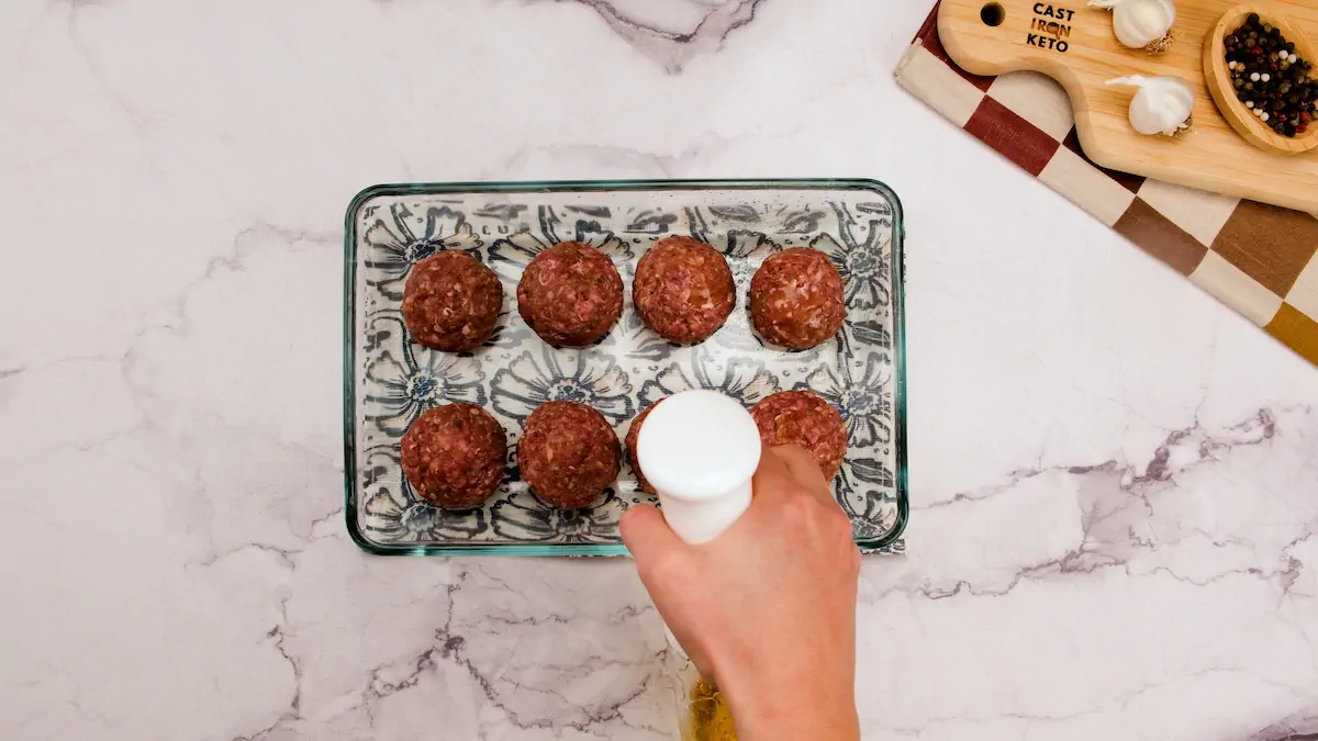 Spraying the meatballs in a rectangular glass bowl with olive oil from a spray bottle.