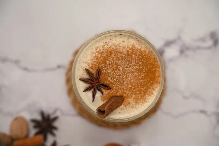 A top-down view of eggnog in a glass, garnished with cinnamon sticks, star anise, and a sprinkle of cinnamon and nutmeg powder.