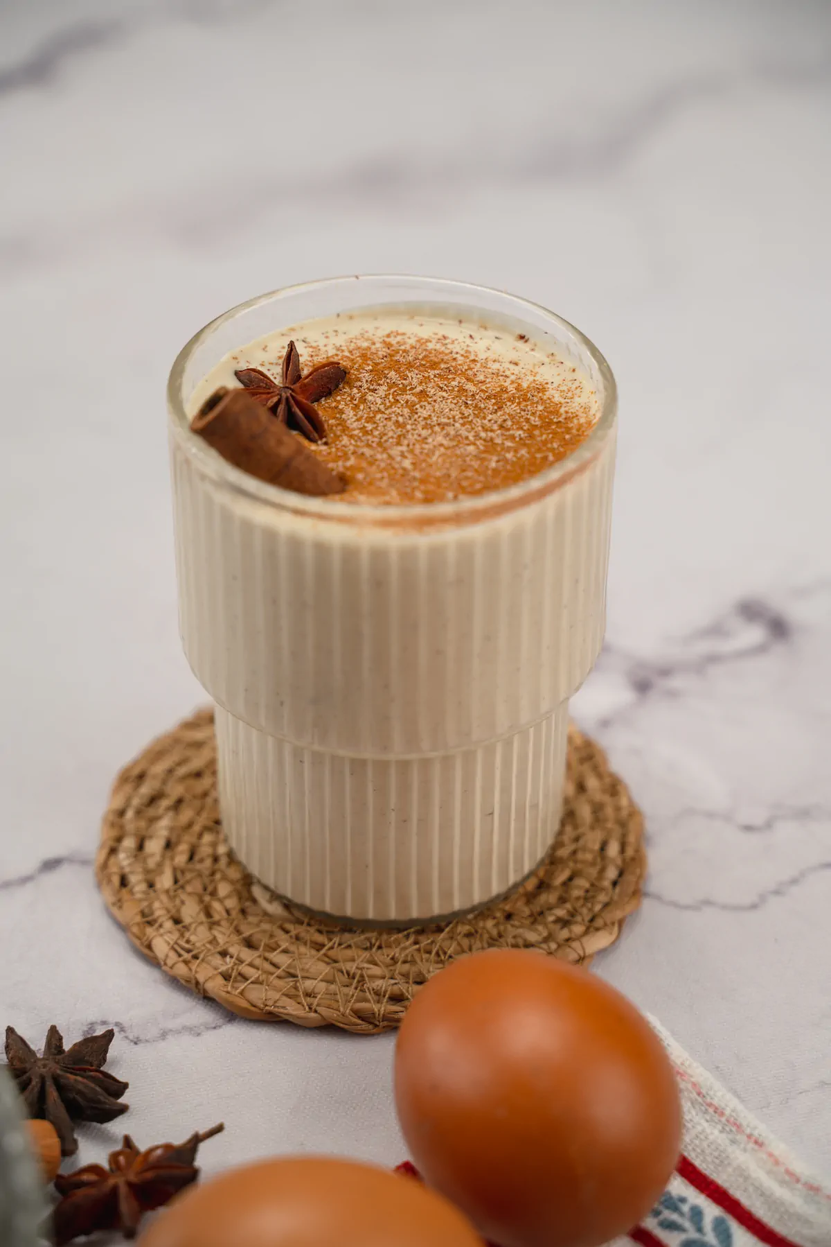 Keto eggnog served in a transparent glass, decorated with cinnamon sticks, star anise, and sprinkled with cinnamon and nutmeg powder.