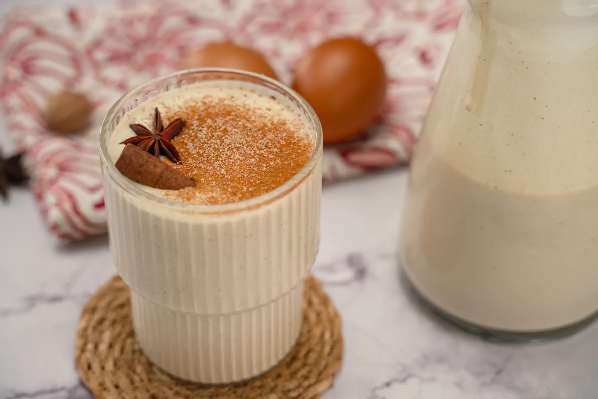 A see-through glass showcasing keto eggnog garnished with powdered spices, cinnamon sticks, and a star anise alongside a glass jar with eggnog.