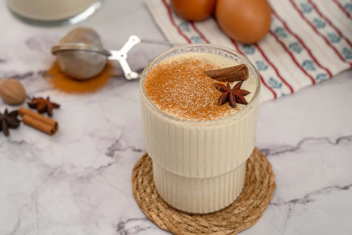 Transparent glass holding keto eggnog with cinnamon sticks, star anise, and a dusting of cinnamon and nutmeg powder.