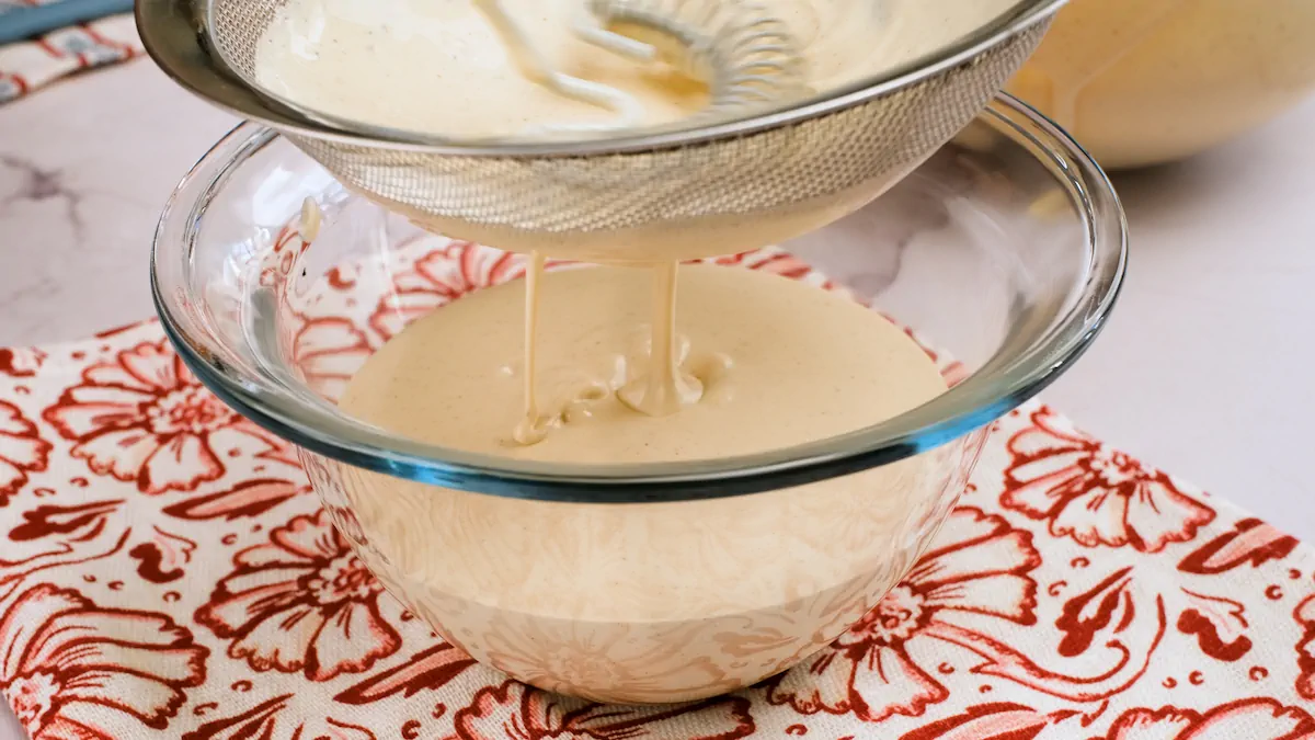 Straining the eggnog mixture into a glass bowl with a stainless steel strainer.