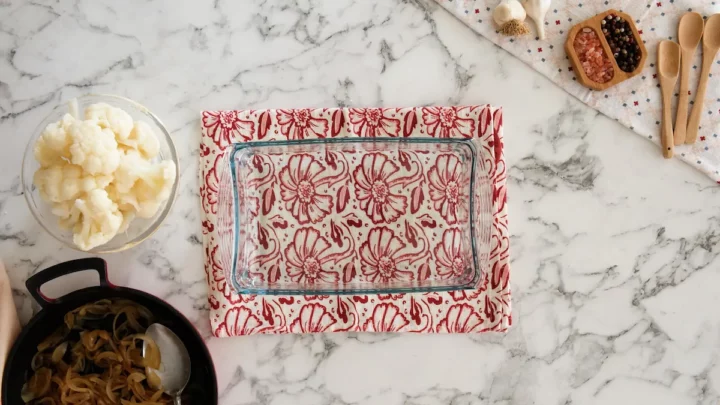 An empty glass baking tray alongside a bowl of blanched cauliflower florets and caramelized onions.