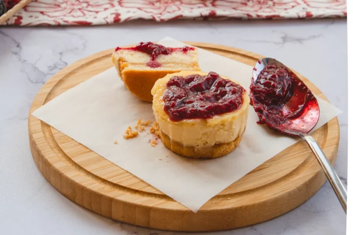 Low-carb mini cheesecake bites topped with raspberry jam, with one cut in half to reveal the texture, alongside a spoonful of the same jam.