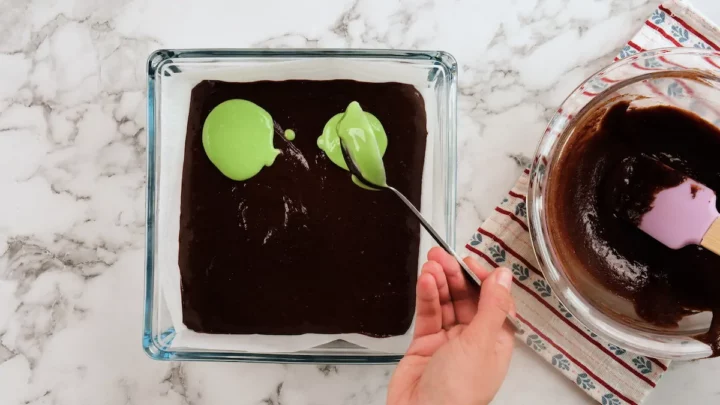 Pouring the green filling with a ladle over the brownie batter in a glass baking dish lined with parchment paper.