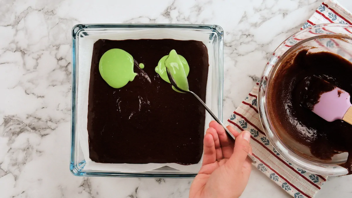 Pouring the green filling with a ladle over the brownie batter in a glass baking dish lined with parchment paper.