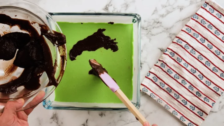 Adding the remaining chocolate brownie batter over the green filling layer with a silicone spatula.